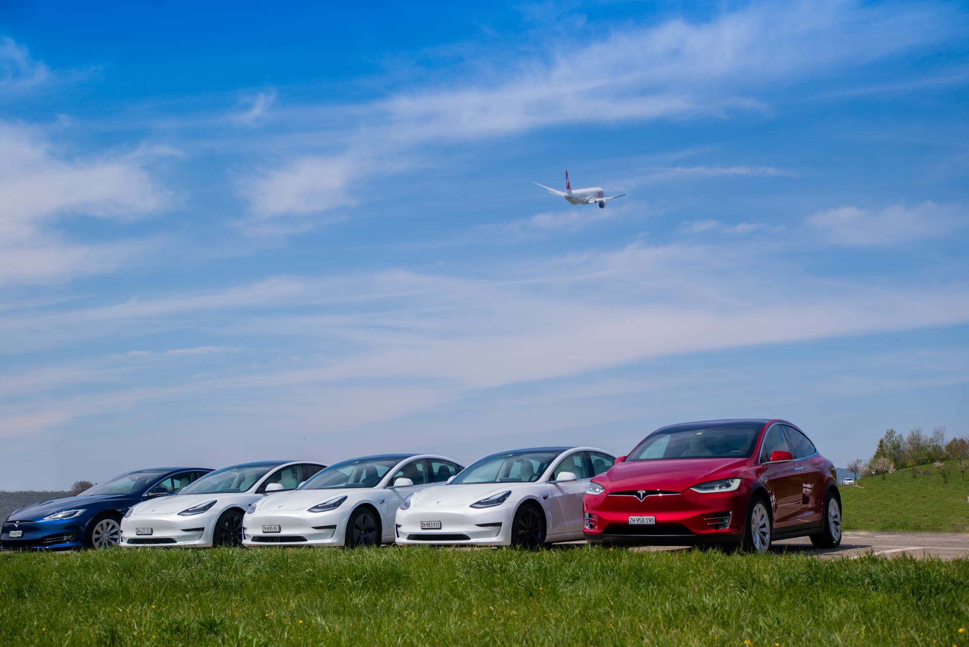 Fünf Teslas von teslify in einer Reihe auf einer Wiese, darüber ein startendes Flugzeug